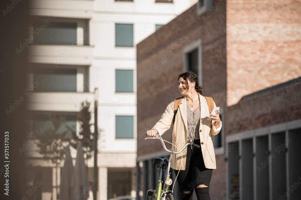 Smiling female student looking away while walking with bicycle against buildings Smiling female student looking away while walking with bicycle against buildings