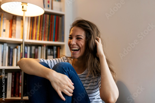 Cheerful young woman with hand in hair laughing while sitting against wall at home photo