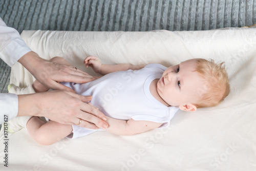 Womens hands do gymnastics for a beautiful baby in a white bodysuit. The baby is lying on the changing table