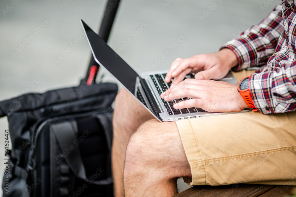 Close up of man hand typing on laptop keyboard while sitting on wooden bench in downtown. The male freelancer uses laptop and an electric scooter. Message, idea. Workspace any ware Close up of man hand typing on laptop keyboard while sitting on wooden bench in downtown. The male freelancer uses laptop and an electric scooter. Message, idea. Workspace any ware