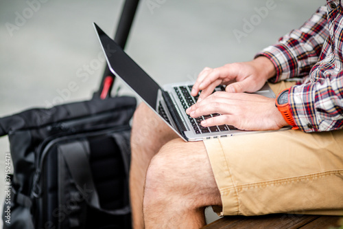 Close up of man hand typing on laptop keyboard while sitting on wooden bench in downtown. The male freelancer uses laptop and an electric scooter. Message, idea. Workspace any ware