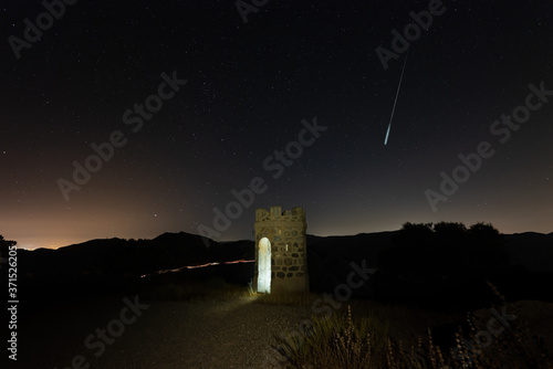 A historical stone watchtower at the Cabo Tinoso fortress. Above it a starry sky with a streak of light from the Perseids. On the horizon the lights of the city of Mazarron. photo