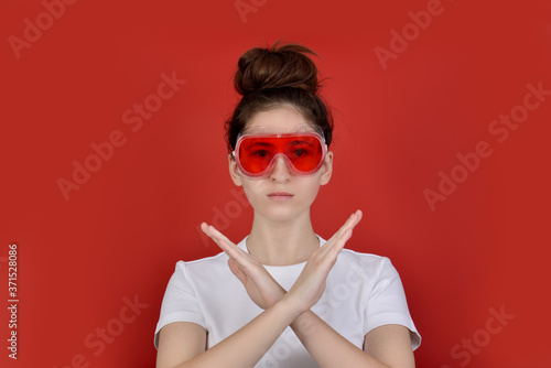 portrait of a serious young brunette girl in a white t shirt and red safety glasses, gesturing no sign, looking at camera, isolated on red background