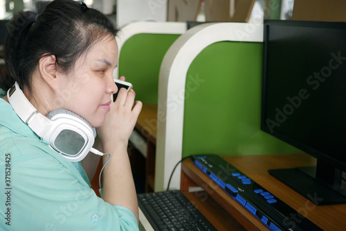 Asian young blind person woman with headphone using smart phone with voice assistive technology for disabilities persons in workplace with computer and braille display on table.