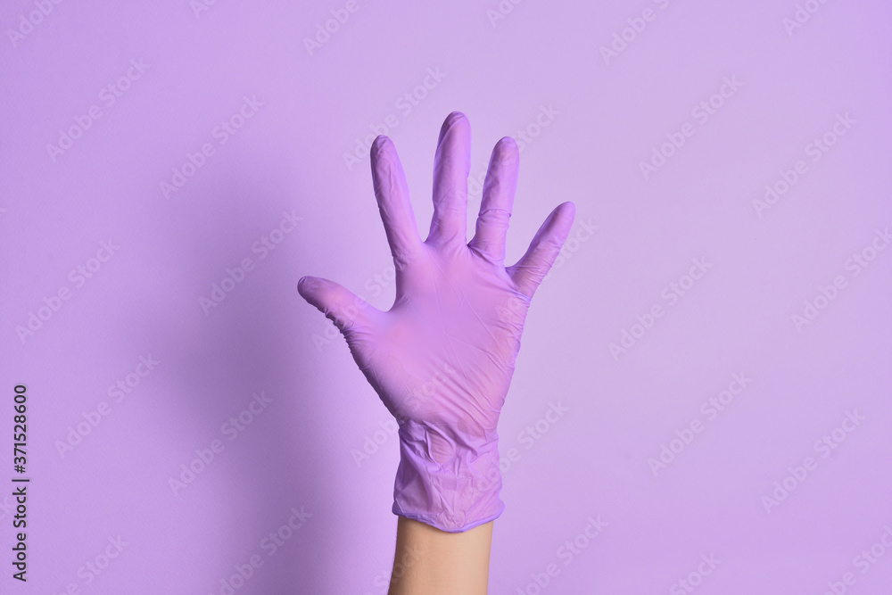 A woman's hand in a lilac-colored medical rubber glove shows an open hand gesture against an isolated purple background. Five fingers