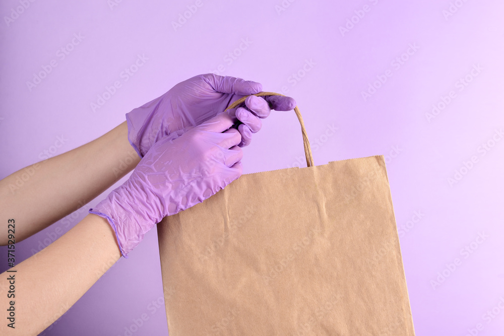 Women's hands in lilac medical gloves hold a paper bag of food on an isolated purple background. Ready-made food delivery Concept Women's hands in lilac medical gloves hold a paper bag of food on an isolated purple background. Ready-made food delivery Concept