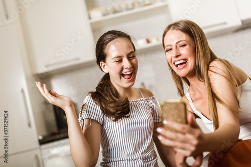 Two joyful cheerful girls taking a selfie while sitting together at home