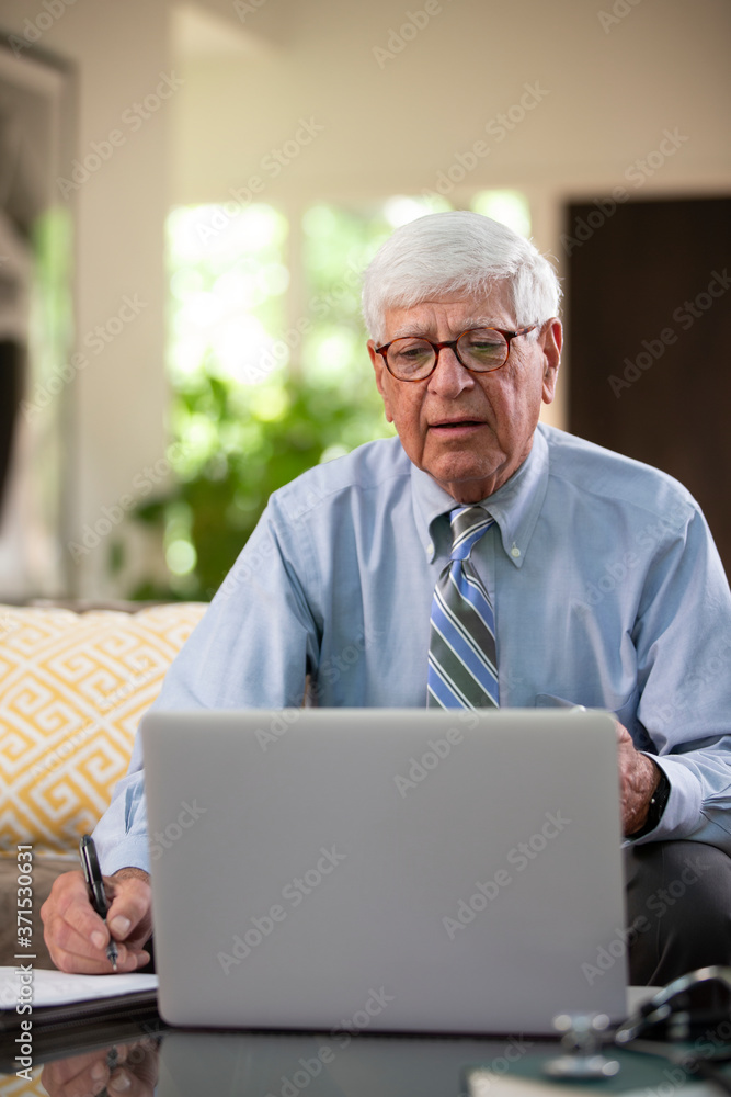 Mature Caucasian Male doctor practicing tele-medicine from his home using laptop computer, Talking to patient and taking notes Mature Caucasian Male doctor practicing tele-medicine from his home using laptop computer, Talking to patient and taking notes