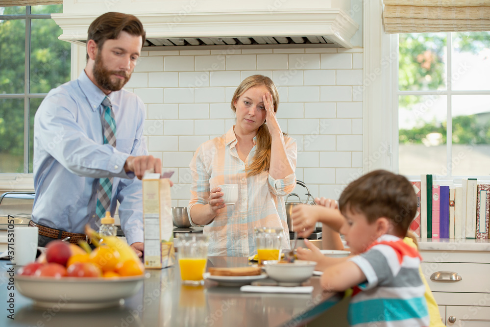 Mom who is feeling under the weather with a headache with family in kitchen, struggling to get the kids breakfast and ready to start the day Mom who is feeling under the weather with a headache with family in kitchen, struggling to get the kids breakfast and ready to start the day