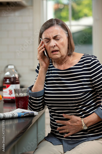 Senior Caucasian woman in her kitchen describing her abdominal pain to doctor on the phone