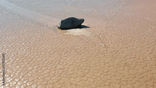 Scenic close up view of rock on the wet cracked surface of Death Valley desert. Epic race track playa park. Sun light reflecting on the slippery dry lake bed. Cinematic 4K aerial, nature background