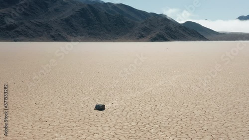 Epic race track playa with moving rocks by the dry cracked surface of Death Valley desert. Cinematic 4K aerial around the moving stones on sunny morning, with mountains and clouds on moving background