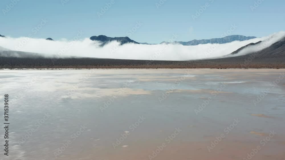 Aerial view of a drone flying over the flat wet cracked surface of Death Valley desert with high black mountains covered by thick fog clouds on background. Sunrise on sunny winter day, 4K California Aerial view of a drone flying over the flat wet cracked surface of Death Valley desert with high black mountains covered by thick fog clouds on background. Sunrise on sunny winter day, 4K California
