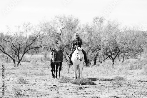 Western ranch lifestyle in black and white, shows woman riding horse bareback while ponying another side by side.