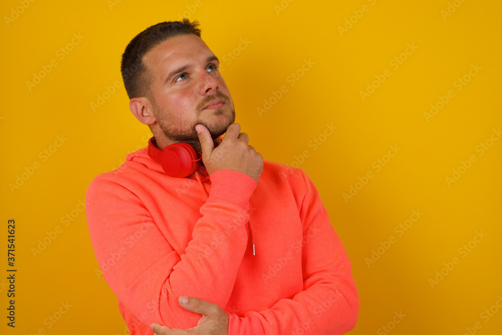 Portrait of thoughtful smiling man keeps hand under chin, looks sideways, thinking or wondering about something with interest, dressed casually, poses against gray studio. Taking decisions concept. Portrait of thoughtful smiling man keeps hand under chin, looks sideways, thinking or wondering about something with interest, dressed casually, poses against gray studio. Taking decisions concept.