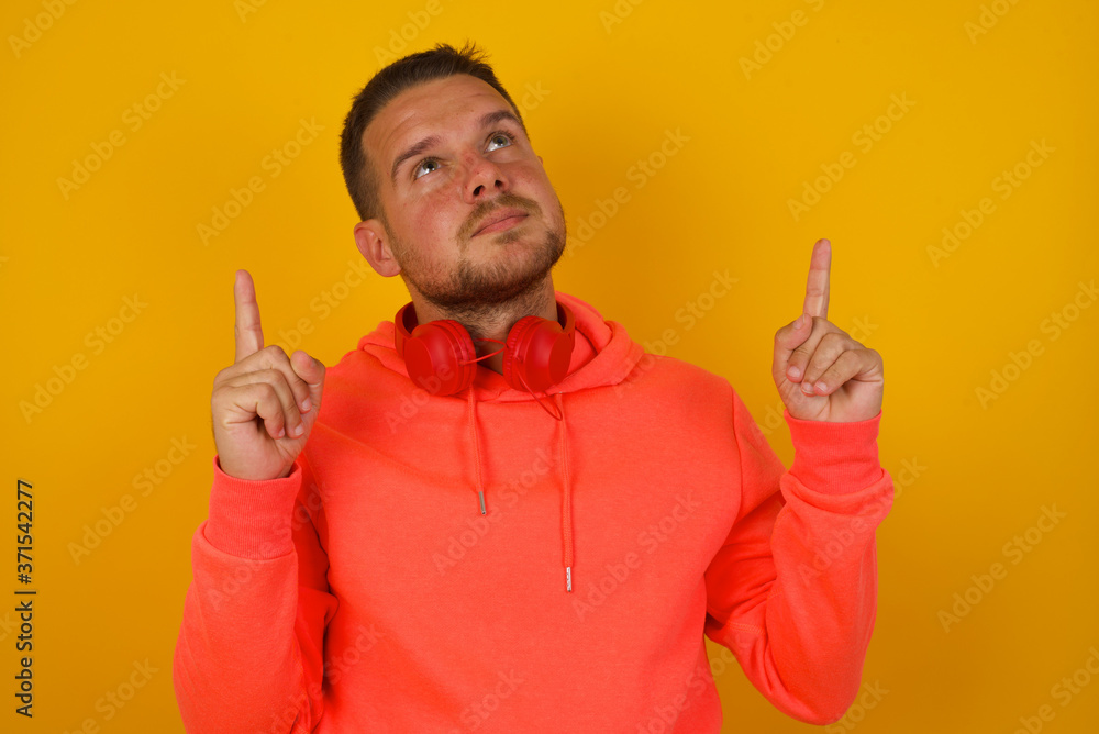 Horizontal shot of successful friendly looking young dark haired man exclaiming excitedly, pointing both index fingers up, indicating something shocking on blank copy space wall.