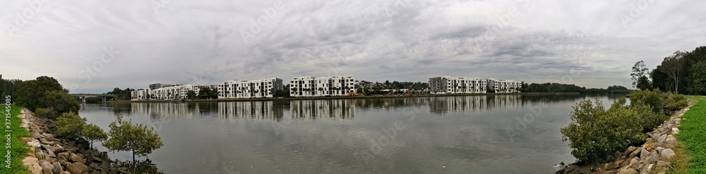Beautiful panoramic view of a river with reflections of modern apartment buildings, cloudy sky and trees on water, Parramatta river, Wilson Park, Silverwater, Sydney, New South Wales, Australia
