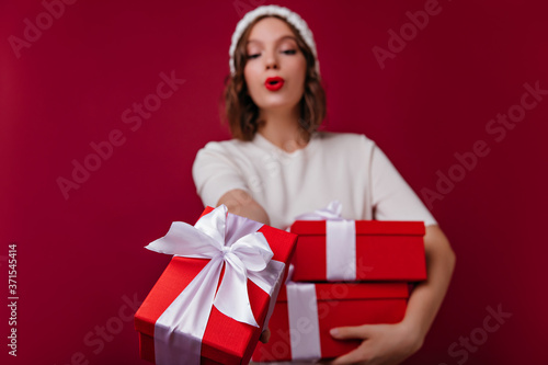 Blur portrait of funny girl enjoying christmas photoshoot. Indoor photo of blissful lady in white hat holding present.