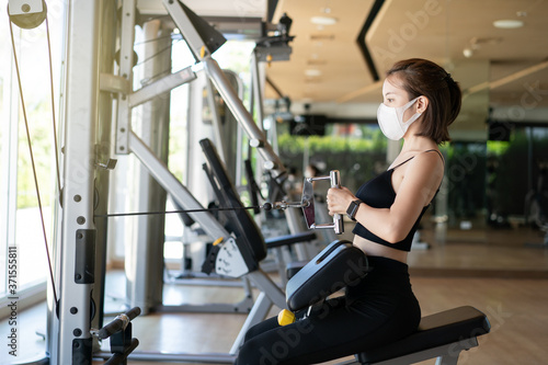 Woman wearing face mask seated cable row, pulling cable of rowing machine training in gym. during corona virus pandermic.