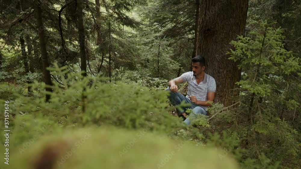 Distant handsome young man leaning on tree is dealing with wood stick in his hand, relaxation concept Distant handsome young man leaning on tree is dealing with wood stick in his hand, relaxation concept