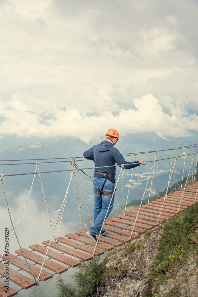 A man and a woman wearing a safety net. They are walking on a rope bridge over an abyss. Crossing a suspension bridge in the mountains. Extreme sport. The view from the back. On a background of clouds