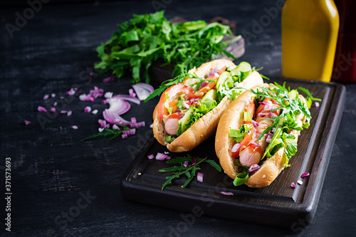 Hot dog with sausage, cucumber, tomato and red onion on dark background.