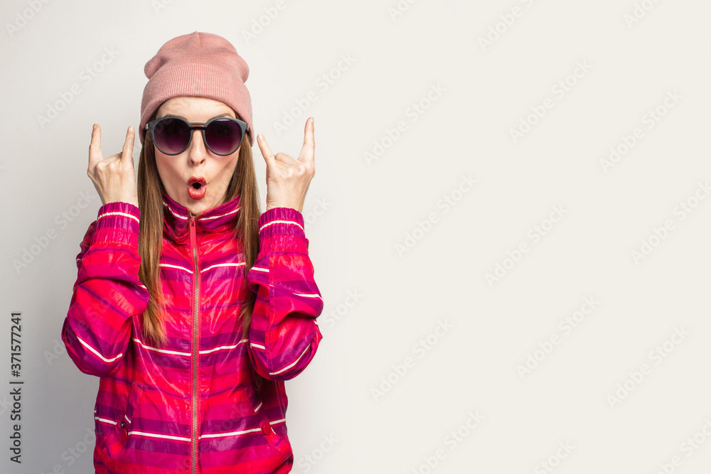 Young woman in a hat and glasses and a pink sports jacket makes a rock and roll goat gesture, we'll catch this party on a white background with empty space. Concept. Face expression. Banner Young woman in a hat and glasses and a pink sports jacket makes a rock and roll goat gesture, we'll catch this party on a white background with empty space. Concept. Face expression. Banner