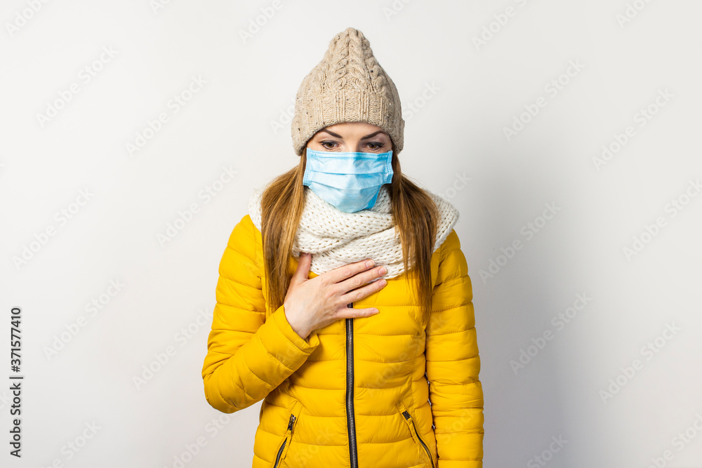 young girl in a yellow jacket and hat with a medical mask on her face holding her hand to her chest and coughing on a light background. Concept of the common cold, virus, infectious diseases young girl in a yellow jacket and hat with a medical mask on her face holding her hand to her chest and coughing on a light background. Concept of the common cold, virus, infectious diseases