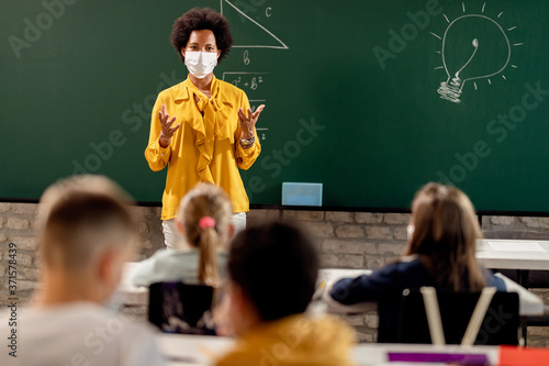 African American elementary teacher wearing face mask while giving a lecture to her students in the classroom.