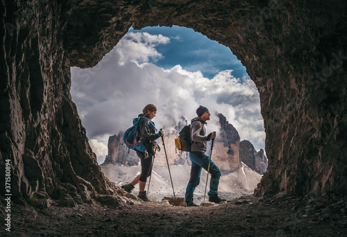 Couple of hikers walking, with Tre Cime di Levarado peaks on background, view from a cave window, in Dolomites, Italy. 