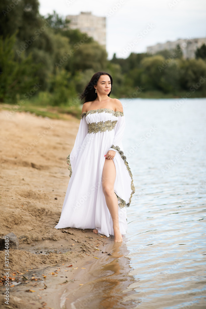 Young european woman with brown hair dressed a long white wedding dress stays near the river Young european woman with brown hair dressed a long white wedding dress stays near the river