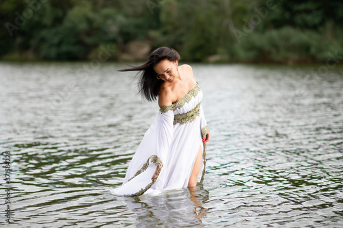 Fototapeta Naklejka Na Ścianę i Meble -  Young european woman with brown hair dressed a long white wedding dress stays in the river