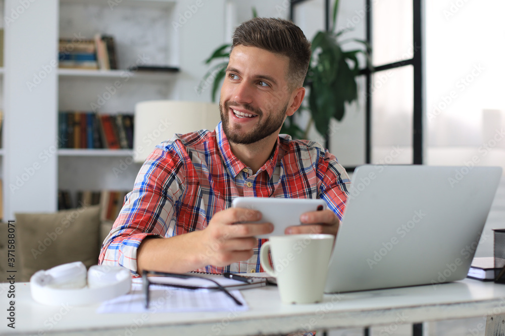 Attractive young man sitting at desk at home office and using mobile phone for cheking social nets. Attractive young man sitting at desk at home office and using mobile phone for cheking social nets.