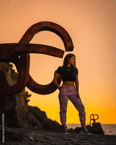 A young pretty brunette Latina with long straight hair in a short black T-shirt and pink pants. Orange sunset next to a famous sculpture of San Sebatian called Peine del Viento, Spain photo