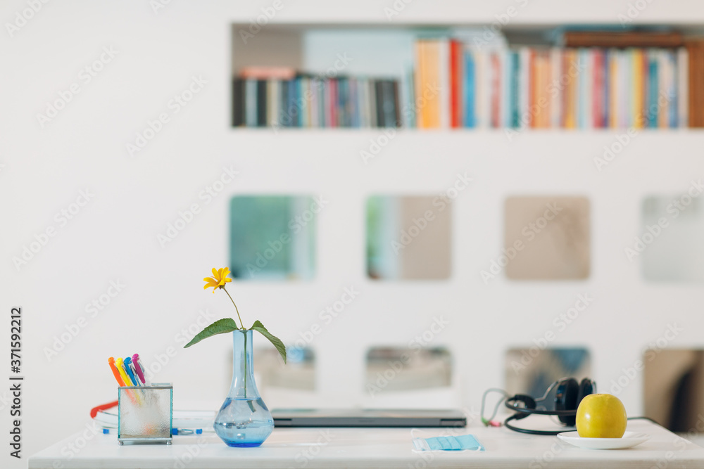 Modern house interior workspace with a silver laptop, headphones, vase, flower, apple, face mask and pens on the table with white wall background and book shelf.