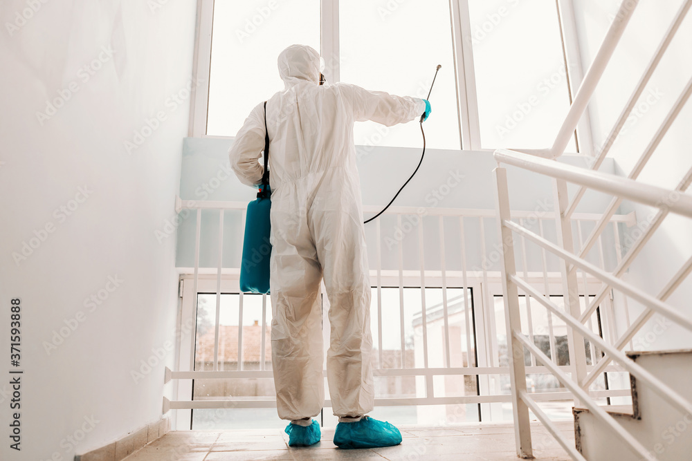 Worker in white sterile uniform, with rubber gloves and mask on holding sprayer with disinfectant and sterilizing windows in school..
