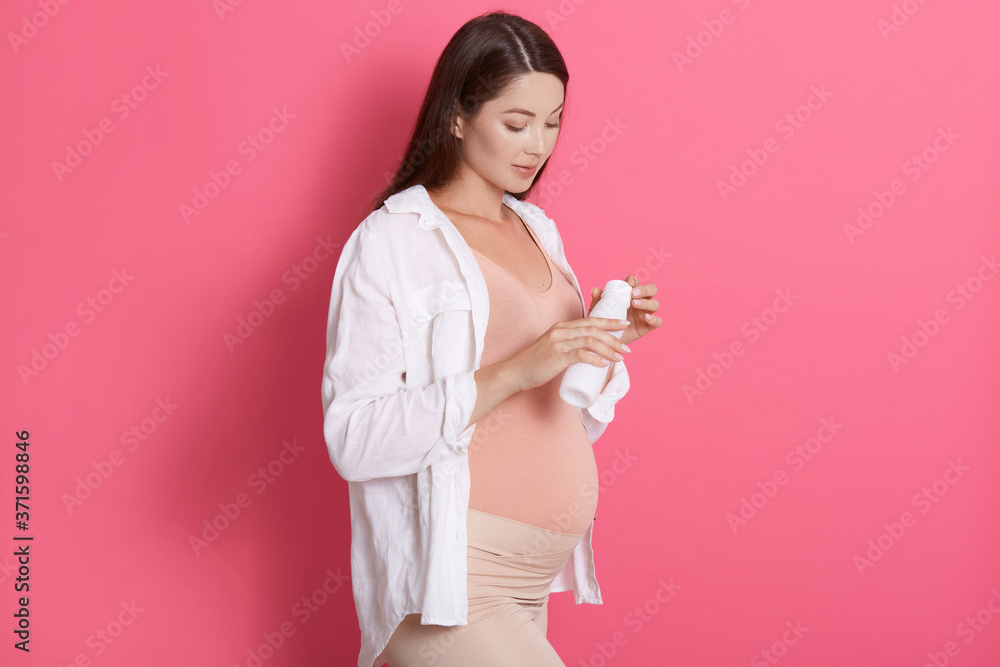 Active healthy pregnant woman holding bottle of milk or yogurt against rosy wall, looking at her beverage, wearing shirt and leggins, having dark straight hair. Active healthy pregnant woman holding bottle of milk or yogurt against rosy wall, looking at her beverage, wearing shirt and leggins, having dark straight hair.