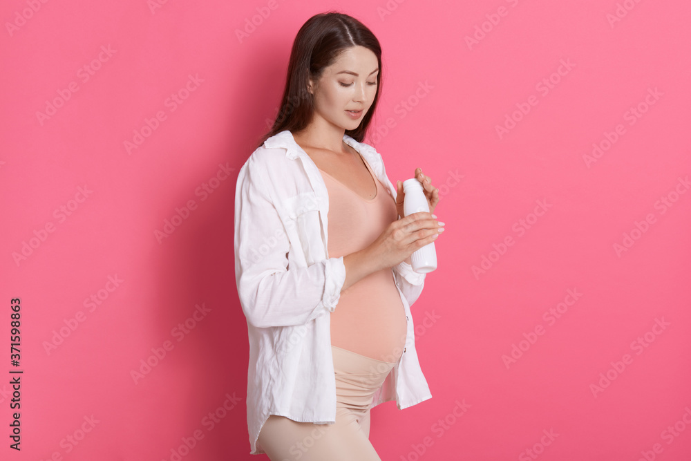 Pregnant woman holding bottle of milk in hands, expectant mother posing isolated over rosy wall, looking at her yogurt, wearing white shirt and leggins. Pregnant woman holding bottle of milk in hands, expectant mother posing isolated over rosy wall, looking at her yogurt, wearing white shirt and leggins.