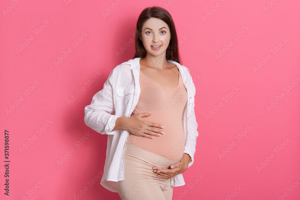Beautiful pregnant woman hugging her tummy and looking at camera with calm happy facial expression, lady with pleasant appearance wearing casual clothing touches her tummy.