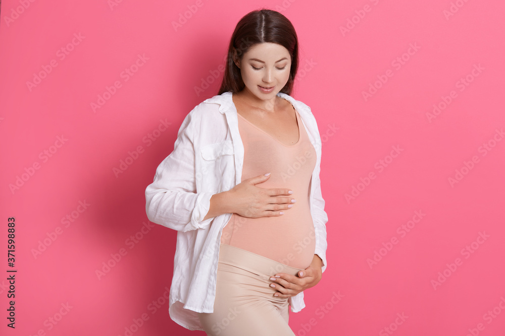 Portrait of beautiful pregnant woman holding her belly and looking at it with great love while posing isolated over rosy background, expectant mommy wearing stylish clothing. Portrait of beautiful pregnant woman holding her belly and looking at it with great love while posing isolated over rosy background, expectant mommy wearing stylish clothing.
