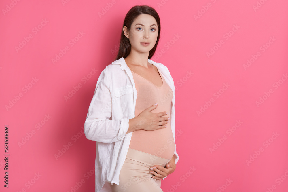 Pregnant woman with hands on stomach looks at camera with serious facial expression, wearing white shirt and leggins, dark haired expectant mother posing indoor. Pregnant woman with hands on stomach looks at camera with serious facial expression, wearing white shirt and leggins, dark haired expectant mother posing indoor.
