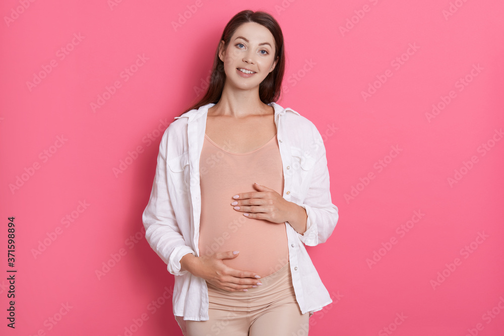 Young beautiful pregnant woman standing isolated over pink background and looking at camera with charming smile. keeping both hands on belly, has dark hair. Young beautiful pregnant woman standing isolated over pink background and looking at camera with charming smile. keeping both hands on belly, has dark hair.