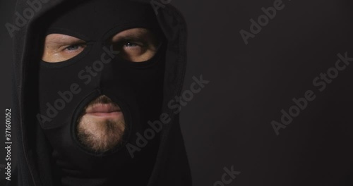 Close up portrait of robber in black clothes and mask looking aggressive to camera on black studio background with smoke. Security system concept.  4K 50 fps slow motion