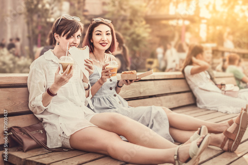 Two happy women friends relax in park and eat street food to take away and drink soft drinks. Casual socializing and urban fast food holiday