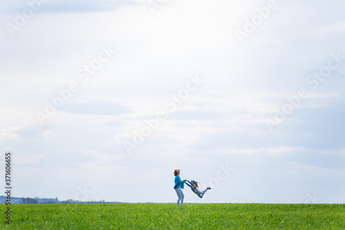 little girl child and mother woman run and jump, green grass in the field, sunny spring weather, smile and joy of the child, blue sky with clouds