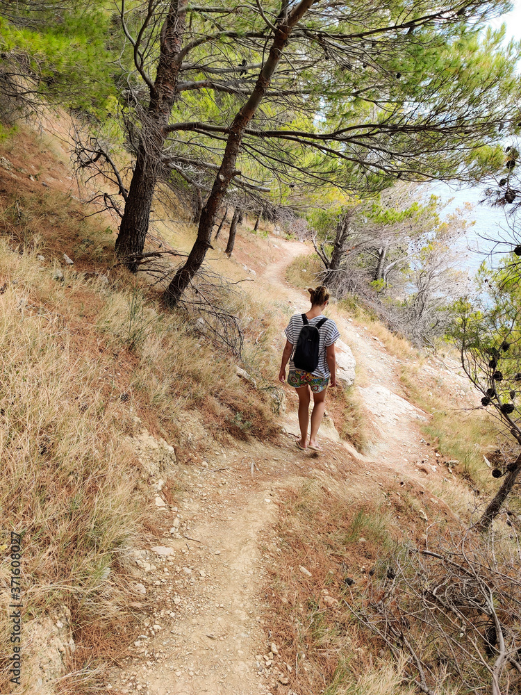 Obraz premium Young active feamle tourist wearing small backpack walking on coastal path among pine trees looking for remote cove to swim alone in peace on seaside in Croatia. Travel and adventure concept.