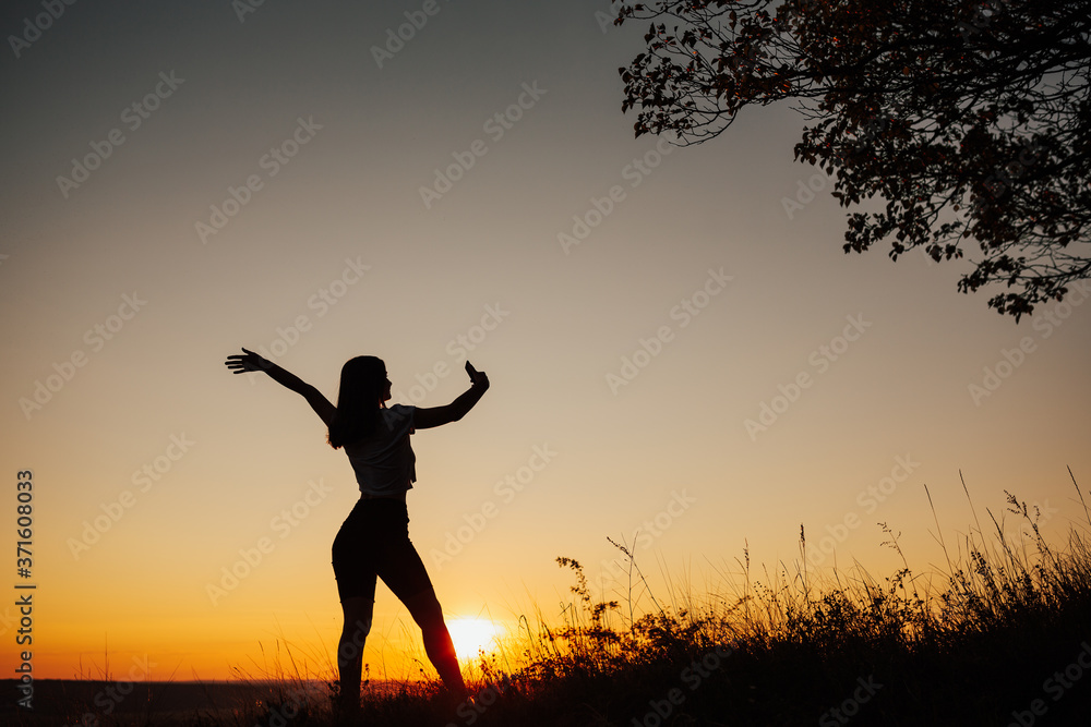 The silhouette of the girl on a background sunset. Happy girl in a field take selfie on a smartphone. Copy space.