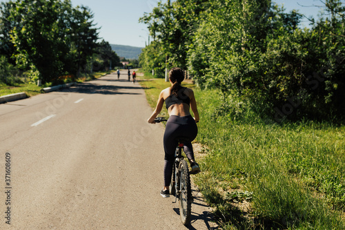 Woman riding bike along asphalt road with green trees on both sides. 