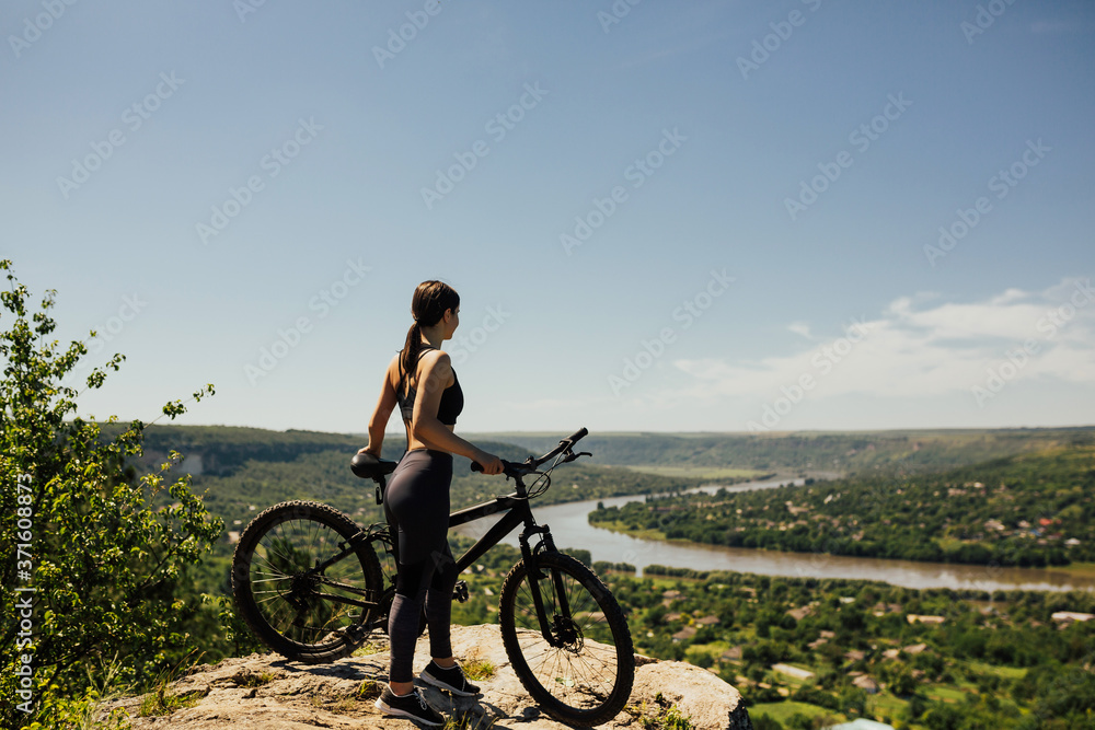 Woman cyclist standing with bicycle on rock on mountain. Young athlete biker with mountain bike on top of the hill observing the river and town view under blue sky.