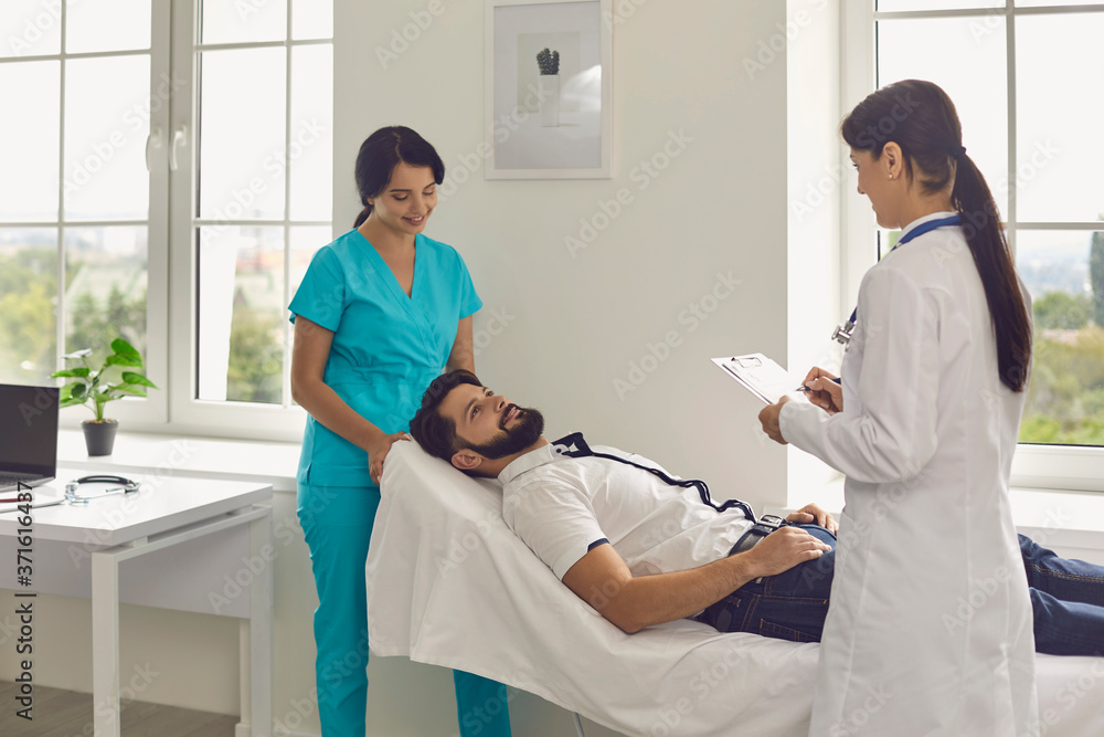 Obraz premium Young doctor and her assistant working with male patient at clinic. Medical specialists helping their client at hospital